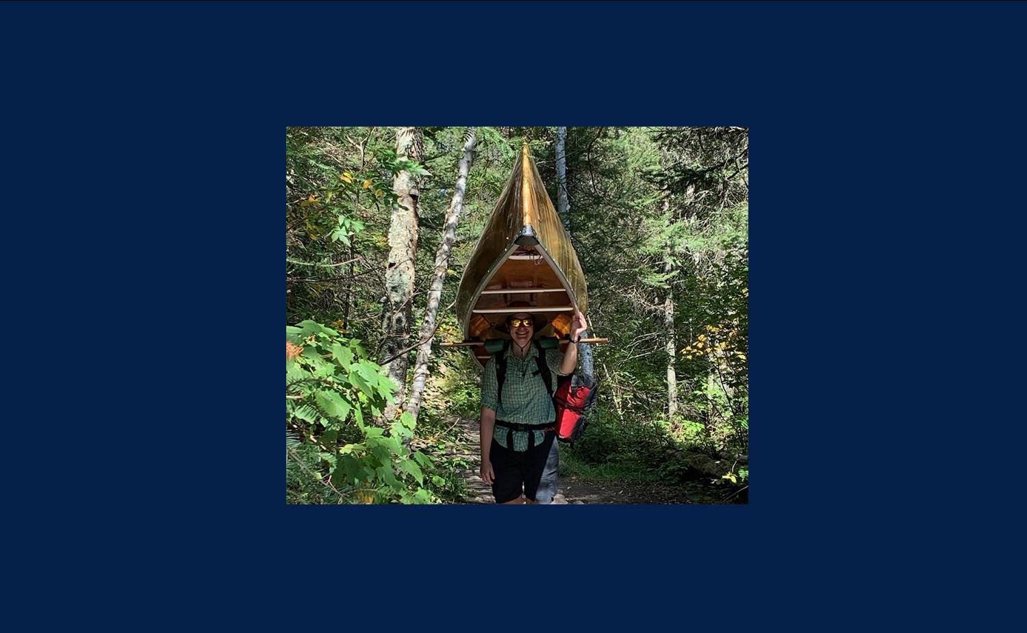 Nina standing in the woods with a canoe on her shoulders